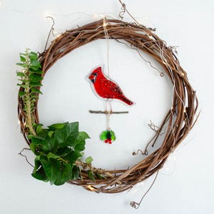 May include: A handcrafted grapevine wreath adorned with a vibrant red cardinal ornament, green ivy, and delicate fairy lights. The cardinal is perched on a small branch, with a sprig of holly below. A festive, nature-inspired decoration.