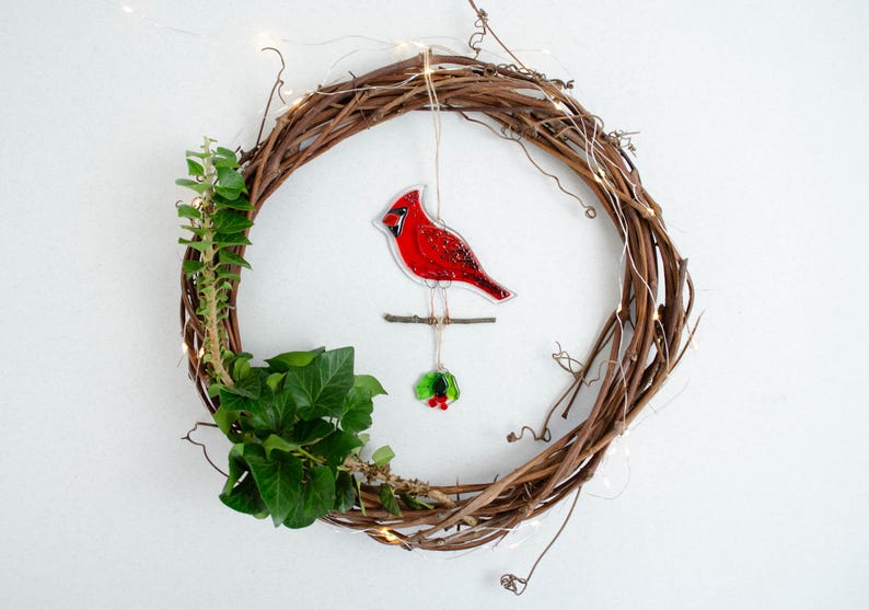 May include: A handcrafted grapevine wreath adorned with a vibrant red cardinal ornament, green ivy, and delicate fairy lights. The cardinal is perched on a small branch, with a sprig of holly below. A festive, nature-inspired decoration.