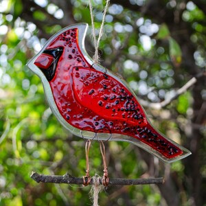 May include: A stained glass cardinal bird decoration, primarily red with black accents, perched on a small branch. The bird is suspended by a string, with copper wire legs. The background is blurred green foliage.
