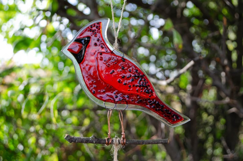 May include: A stained glass cardinal bird decoration, primarily red with black accents, perched on a small branch. The bird is suspended by a string, with copper wire legs. The background is blurred green foliage.