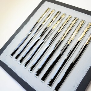 May include: A square wall art piece with a gray background and 10 black and gold glass rods arranged in a vertical pattern.