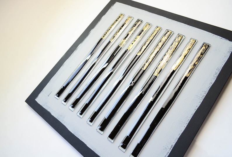 May include: A square wall art piece with a gray background and 10 black and gold glass rods arranged in a vertical pattern.