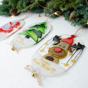 Könnte beinhalten: Drei Weihnachtsbaumschmuck aus Glas mit festlichen Designs. Einer zeigt ein Rentier mit roter Mütze, ein anderer einen grünen Weihnachtsbaum und der dritte einen roten Weihnachtsmann. Jeder Schmuck hat eine Schnur zum Aufhängen und die Worte "Happy New Year!!!" und "Merry Christmas!" sind darauf geschrieben.