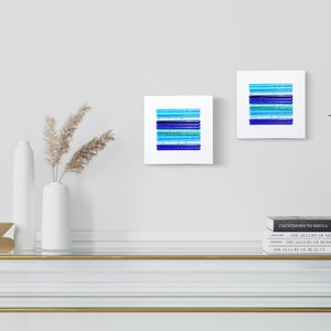 May include: Two square abstract art prints with blue and turquoise stripes on a white background. The prints are hanging on a white wall above a glass and gold console table with a vase of dried pampas grass and books.