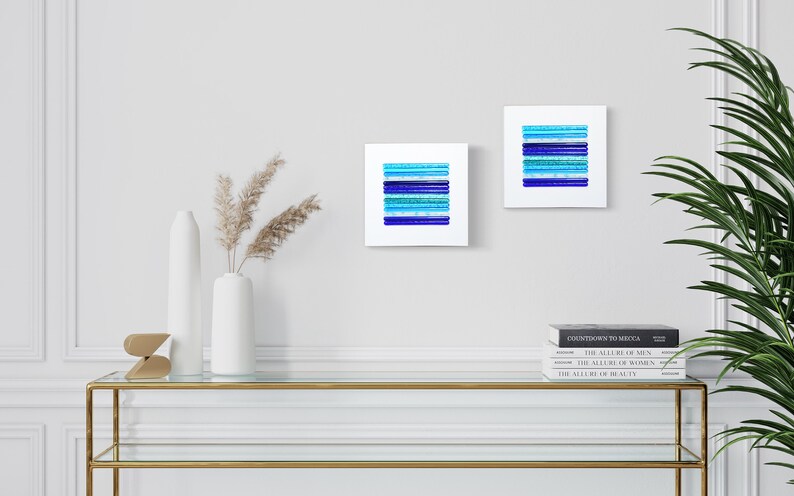 May include: Two square abstract art prints with blue and turquoise stripes on a white background. The prints are hanging on a white wall above a glass and gold console table with a vase of dried pampas grass and books.