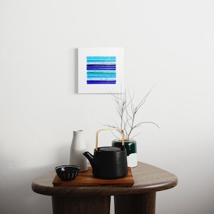 May include: A black teapot with a wooden handle sits on a wooden table with a small bowl and two white vases. A framed abstract painting with blue and green stripes hangs on the wall behind the table.