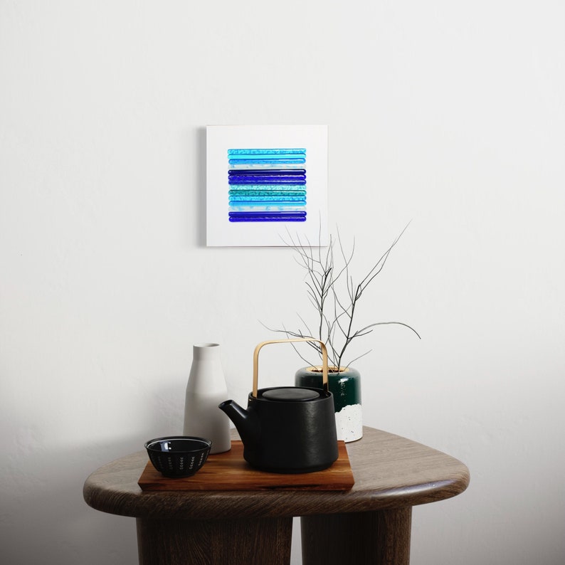 May include: A black teapot with a wooden handle sits on a wooden table with a small bowl and two white vases. A framed abstract painting with blue and green stripes hangs on the wall behind the table.