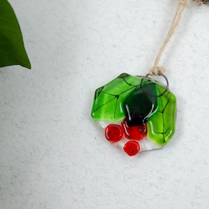 May include: A handmade glass pendant featuring a green leaf design with red berry accents. The pendant is suspended from a natural fiber cord. The glass has a glossy finish and is set against a textured white background.