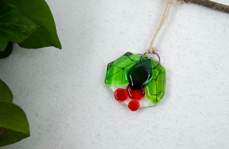 May include: A handmade glass pendant featuring a green leaf design with red berry accents. The pendant is suspended from a natural fiber cord. The glass has a glossy finish and is set against a textured white background.