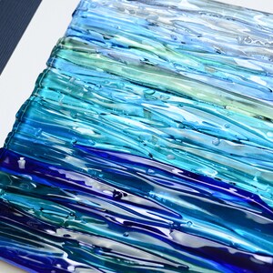 May include: Abstract wall art made of fused glass in shades of blue, teal, and green. The glass is arranged in horizontal stripes, creating a wave-like effect.