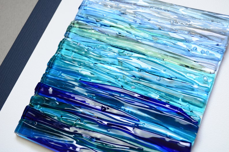 May include: Abstract wall art made of fused glass in shades of blue, teal, and green. The glass is arranged in horizontal stripes, creating a wave-like effect.
