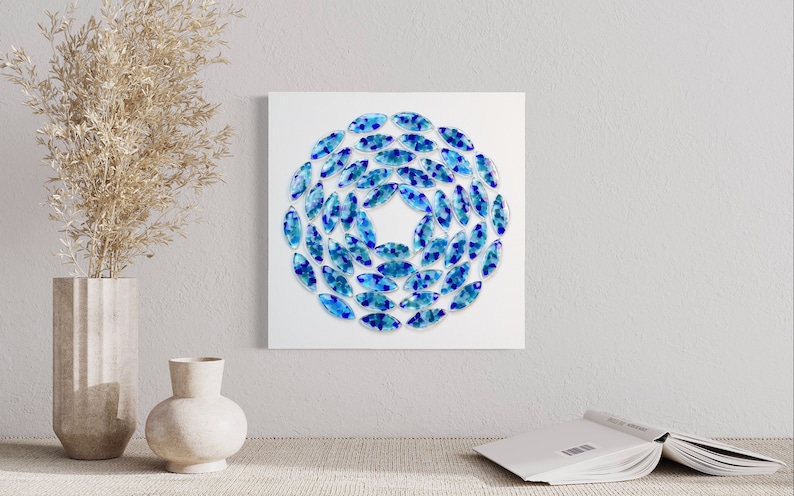 May include: A white square canvas with a blue and teal circular design made of small, oval-shaped glass pieces.