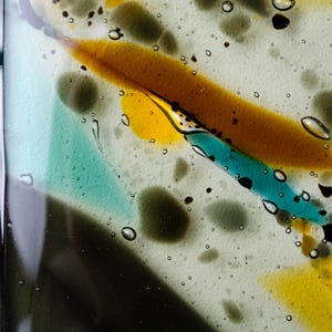 May include: A piece of fused glass featuring a clear base with embedded abstract shapes in shades of amber, turquoise, and dark gray. The surface contains numerous small air bubbles trapped within the glass, creating a textured appearance.