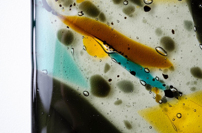 May include: A piece of fused glass featuring a clear base with embedded abstract shapes in shades of amber, turquoise, and dark gray. The surface contains numerous small air bubbles trapped within the glass, creating a textured appearance.