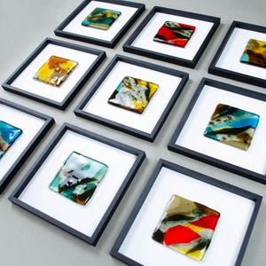 May include: A collection of nine square wall art pieces, each featuring a unique fused glass tile with abstract patterns in various colors including red, yellow, blue, green, and brown, mounted in a black frame with a white mat border.