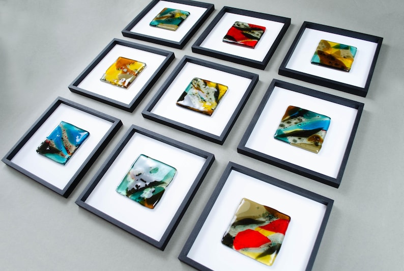 May include: A collection of nine square wall art pieces, each featuring a unique fused glass tile with abstract patterns in various colors including red, yellow, blue, green, and brown, mounted in a black frame with a white mat border.