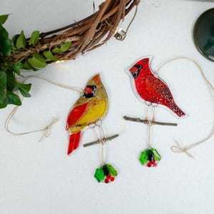 May include: Two stained glass cardinal ornaments, one yellow and red, the other all red, perched on small branches. Each bird hangs from a string, with a sprig of holly below. A grapevine wreath and greenery are in the background.