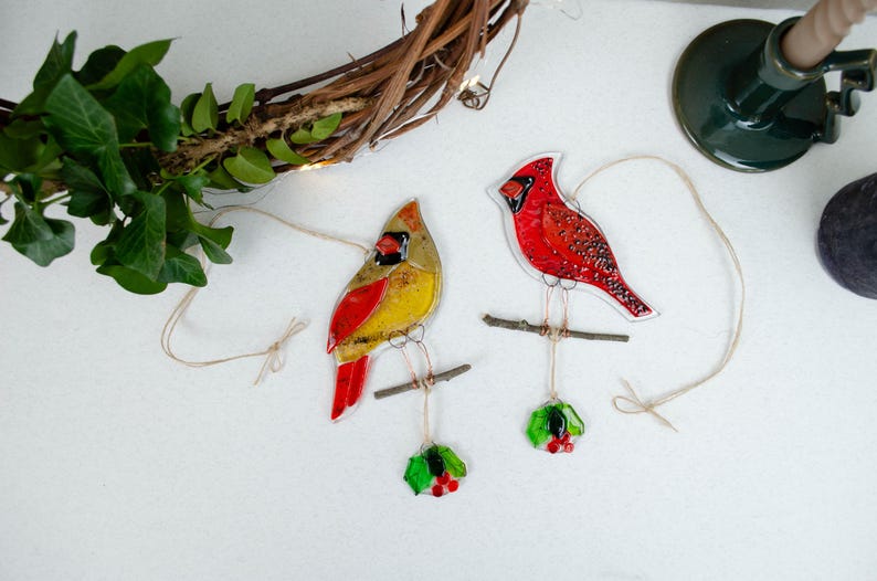 May include: Two stained glass cardinal ornaments, one yellow and red, the other all red, perched on small branches. Each bird hangs from a string, with a sprig of holly below. A grapevine wreath and greenery are in the background.