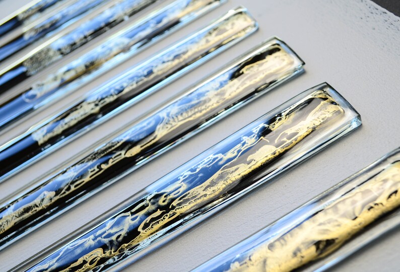 May include: A set of six clear glass rectangular tiles with black and gold abstract designs inside. The tiles are arranged in a row, with the top tile slightly offset from the others.