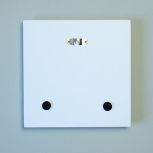 May include: A white square wooden panel with a metal sawtooth hanger and two black circular pads on the back. The panel is designed for wall mounting and has a clean, minimalist aesthetic.