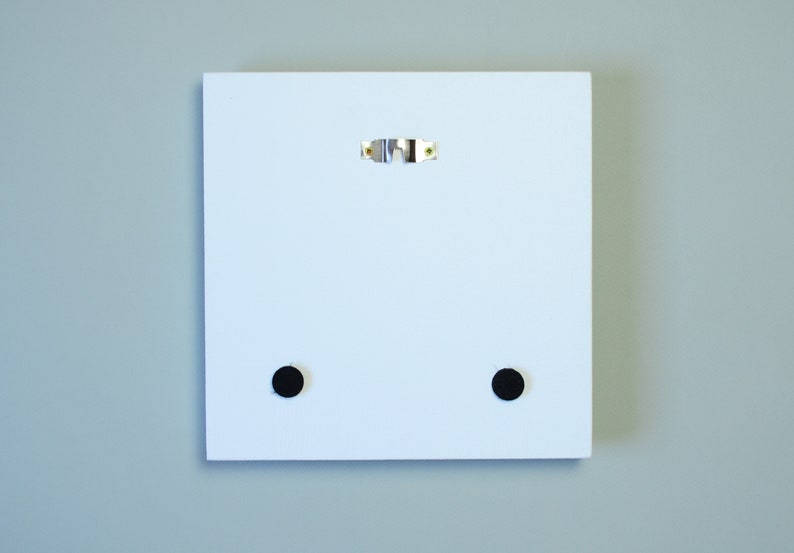 May include: A white square wooden panel with a metal sawtooth hanger and two black circular pads on the back. The panel is designed for wall mounting and has a clean, minimalist aesthetic.
