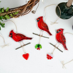 May include: Three red cardinal ornaments with black accents, perched on small branches, each suspended by twine. One ornament has a red heart, another has a holly sprig, and the third has a red and white ornament. The ornaments are on a white surface.