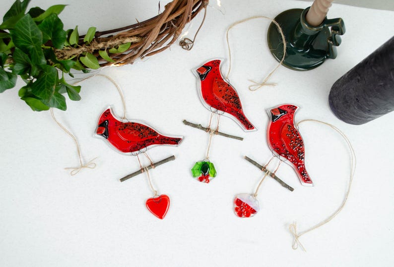 May include: Three red cardinal ornaments with black accents, perched on small branches, each suspended by twine. One ornament has a red heart, another has a holly sprig, and the third has a red and white ornament. The ornaments are on a white surface.
