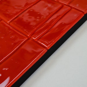May include: Close-up of a section of red glossy tiles arranged in a brick pattern. The tiles are rectangular and have a reflective surface. The tiles are set against a black border and a white background.
