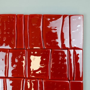 May include: A close-up view of a section of a wall or artwork featuring glossy, rectangular red tiles. The tiles are arranged in a grid-like pattern, with minor variations in shape and size. The background is a light grey colour.