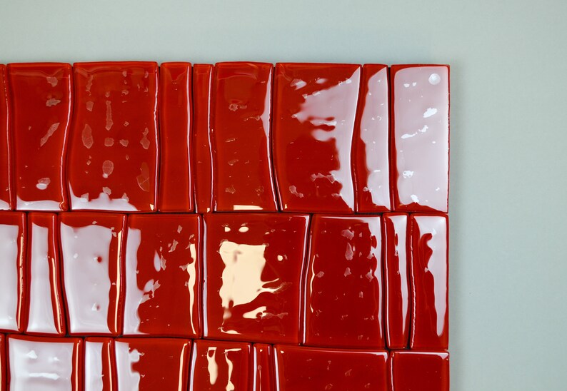 May include: A close-up view of a section of a wall or artwork featuring glossy, rectangular red tiles. The tiles are arranged in a grid-like pattern, with minor variations in shape and size. The background is a light grey colour.
