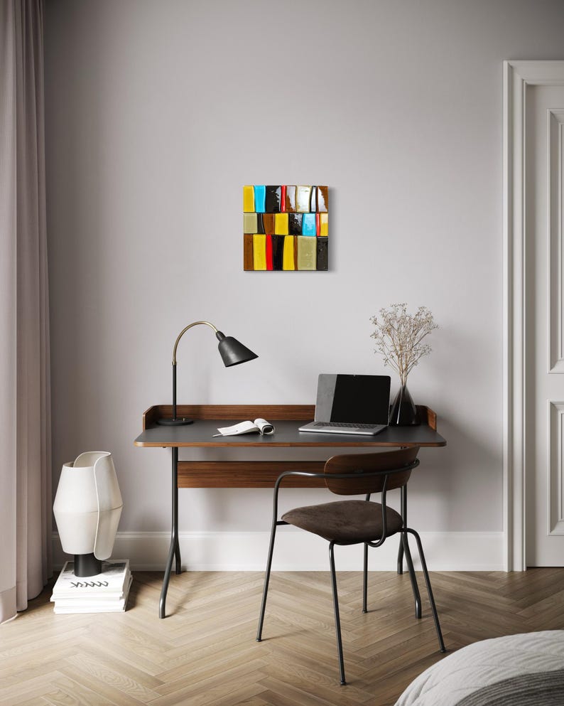 May include: An abstract artwork with a rectangular design, featuring a grid of colourful blocks. The blocks include shades of yellow, blue, red, and brown. The artwork hangs above a desk with a laptop, lamp, and chair, creating a modern office setting.