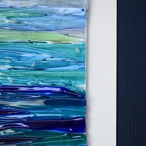May include: Abstract wall art featuring a layered design of blue, green, and purple glass. The glass pieces are arranged in horizontal stripes, creating a wave-like effect. The art is framed in a dark blue wood frame.