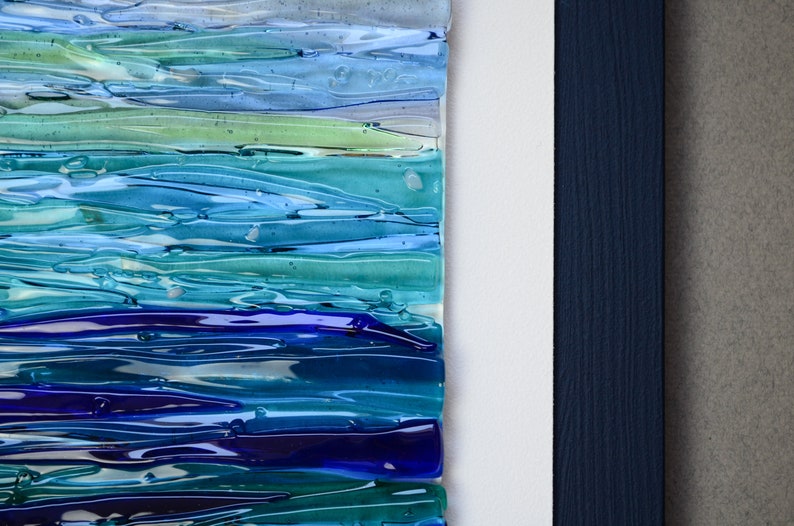 May include: Abstract wall art featuring a layered design of blue, green, and purple glass. The glass pieces are arranged in horizontal stripes, creating a wave-like effect. The art is framed in a dark blue wood frame.