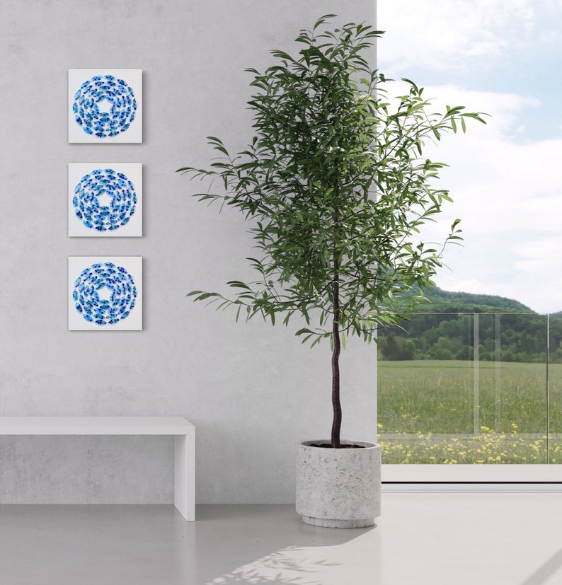 May include: Three framed blue and white abstract paintings hang on a white wall, with a potted green plant in front of a window.