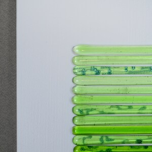 May include: A wall art piece made of green glass tubes arranged in a vertical line. The tubes are of varying lengths and have a slightly textured surface.