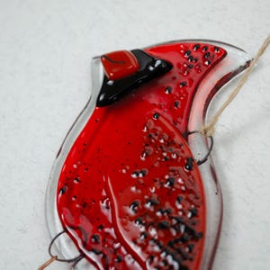 May include: A handcrafted glass cardinal ornament. The bird features a vibrant red body with black accents and a clear glass outline. A natural twine loop is attached for hanging. The ornament is a decorative item.