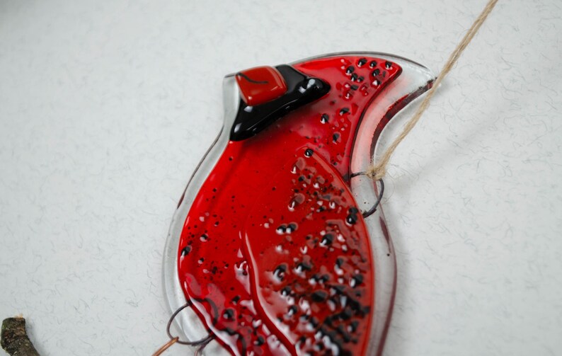 May include: A handcrafted glass cardinal ornament. The bird features a vibrant red body with black accents and a clear glass outline. A natural twine loop is attached for hanging. The ornament is a decorative item.