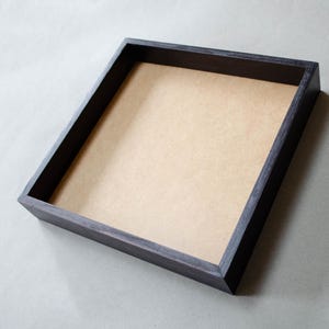 May include: A square wooden tray featuring a dark, stained finish on the outer frame and a flat, light tan interior base. The tray has four straight sides with visible wood grain texture and sharp, clean corners, forming a simple open container.