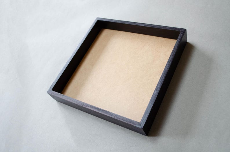 May include: A square wooden tray featuring a dark, stained finish on the outer frame and a flat, light tan interior base. The tray has four straight sides with visible wood grain texture and sharp, clean corners, forming a simple open container.