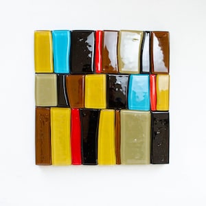 May include: A square mosaic made of rectangular glass tiles in a grid pattern. The tiles are in various colours: yellow, blue, black, red, and brown. The mosaic is set against a white background. The overall effect is a vibrant, abstract design.
