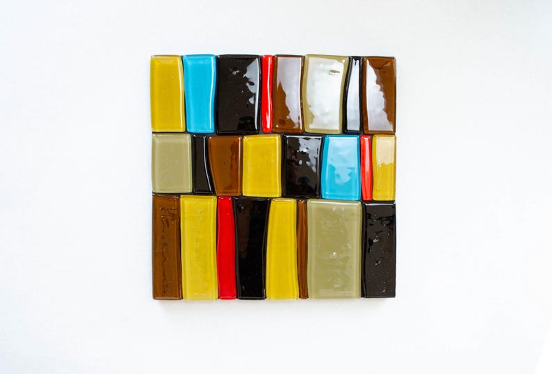 May include: A square mosaic made of rectangular glass tiles in a grid pattern. The tiles are in various colours: yellow, blue, black, red, and brown. The mosaic is set against a white background. The overall effect is a vibrant, abstract design.