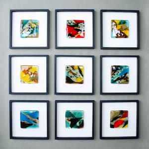 May include: A grid of nine square wall art pieces, each featuring a small, multicolored fused glass tile centered within a white mat and a thin black frame. The glass tiles display various abstract patterns in shades of blue, red, yellow, and green.