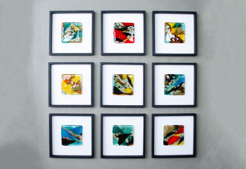 May include: A grid of nine square wall art pieces, each featuring a small, multicolored fused glass tile centered within a white mat and a thin black frame. The glass tiles display various abstract patterns in shades of blue, red, yellow, and green.