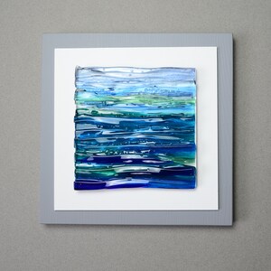 May include: Abstract art piece featuring layered glass in shades of blue and green, resembling a seascape. The artwork is mounted on a white background within a gray frame, creating a modern and serene aesthetic. The piece is square in shape.