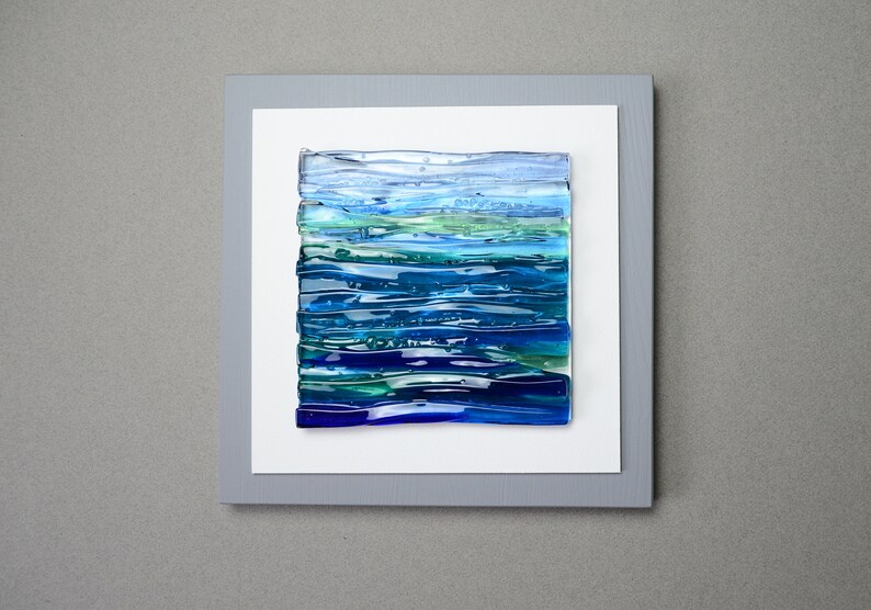 May include: Abstract art piece featuring layered glass in shades of blue and green, resembling a seascape. The artwork is mounted on a white background within a gray frame, creating a modern and serene aesthetic. The piece is square in shape.