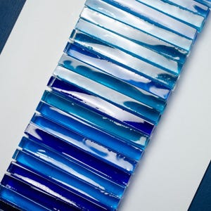 May include: A long strip of blue glass tiles arranged in a gradient pattern, starting with a light blue and transitioning to a dark blue. The tiles are arranged vertically and are set against a white background.