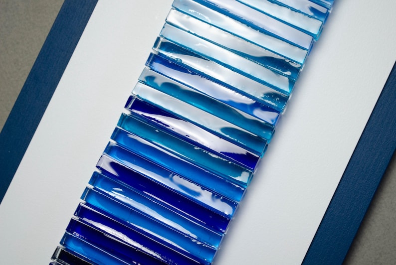 May include: A long strip of blue glass tiles arranged in a gradient pattern, starting with a light blue and transitioning to a dark blue. The tiles are arranged vertically and are set against a white background.