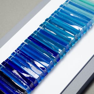 May include: A wall art piece featuring a gradient of blue glass tiles arranged in a vertical line. The tiles are arranged in a graduated pattern, starting with a light blue at the top and transitioning to a dark blue at the bottom.