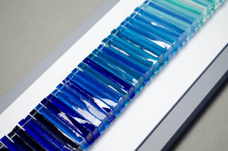 May include: A wall art piece featuring a gradient of blue glass tiles arranged in a vertical line. The tiles are arranged in a graduated pattern, starting with a light blue at the top and transitioning to a dark blue at the bottom.