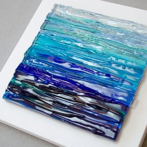 May include: Abstract glass art piece featuring a gradient of blues and greens, resembling ocean waves. The textured glass is mounted on a white square canvas, creating a striking contrast. The artwork evokes a sense of depth and movement.